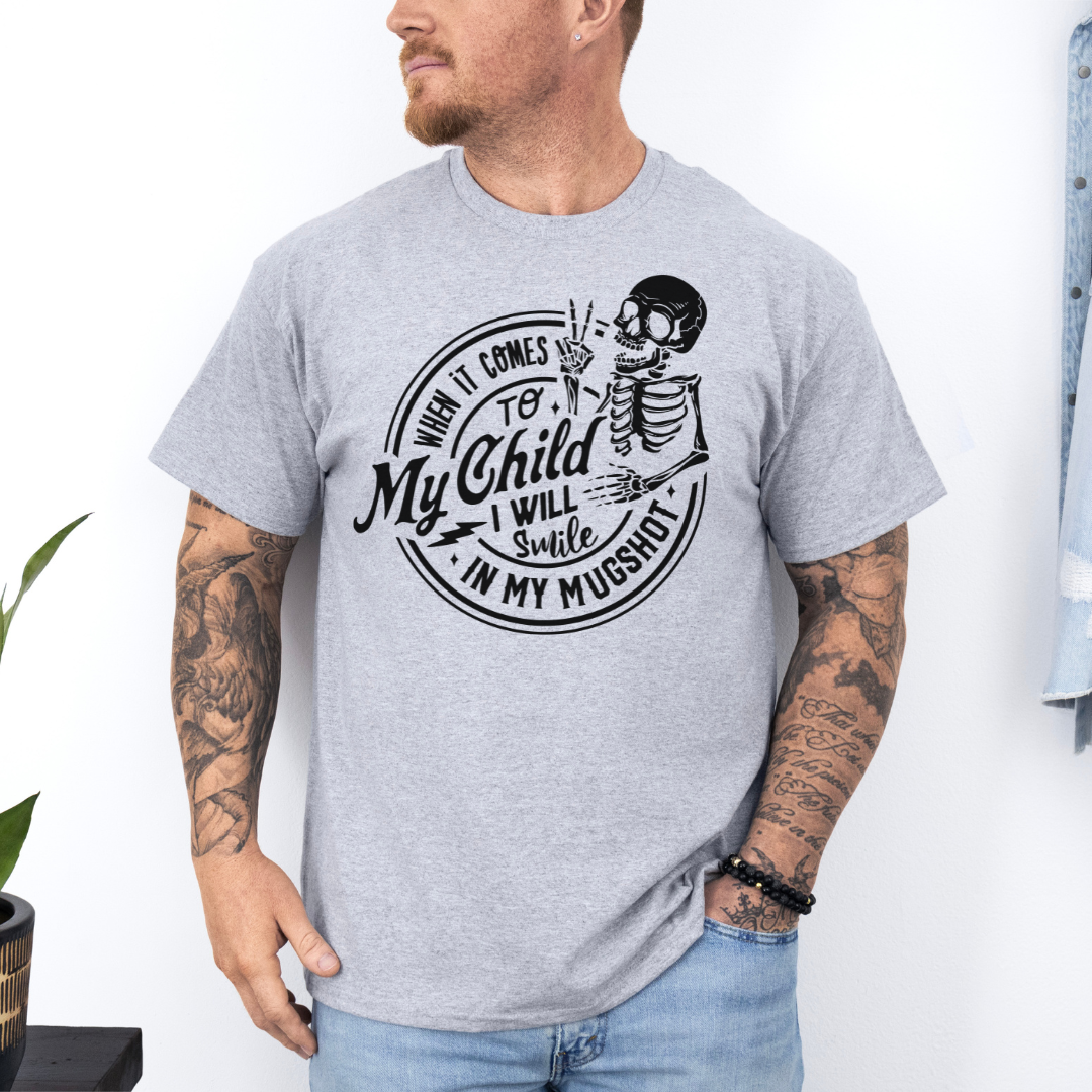 A man wearing a grey graphic t-shirt with a white skeleton and text design Saying "when it comes to my child, i will smile in my mugshot" styled with blue denim jeans. Purchase from Coffee and Moo NZ.