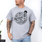 A man wearing a grey graphic t-shirt with a white skeleton and text design Saying "when it comes to my child, i will smile in my mugshot" styled with blue denim jeans. Purchase from Coffee and Moo NZ.
