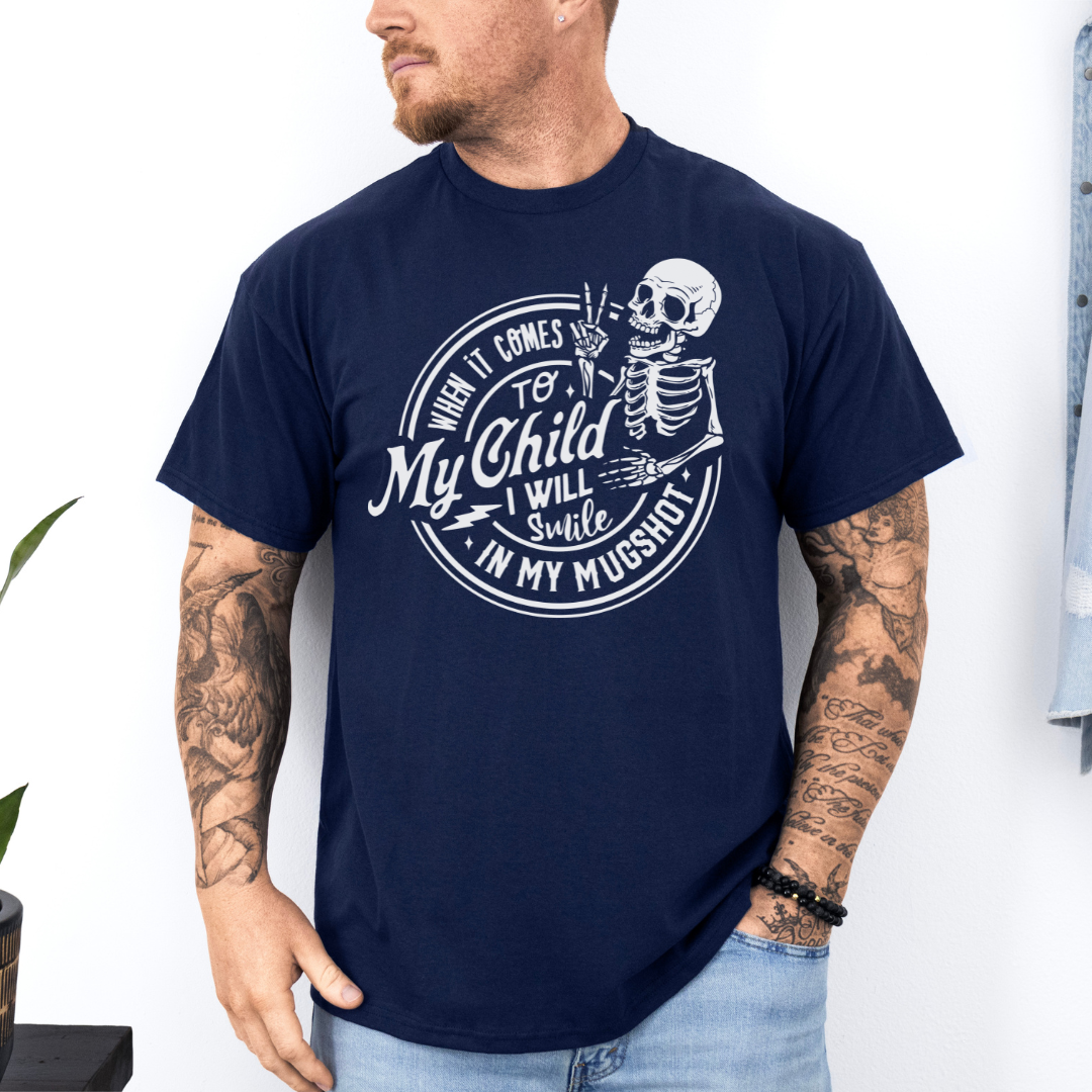 A man wearing a navy graphic t-shirt with a white skeleton and text design Saying "when it comes to my child, i will smile in my mugshot" styled with blue denim jeans. Purchase from Coffee and Moo NZ.