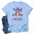 A light blue graphic t-shirt with a cartoon unicorn and profane text, styled flat-lay next to a pair of blue jeans on a white background.