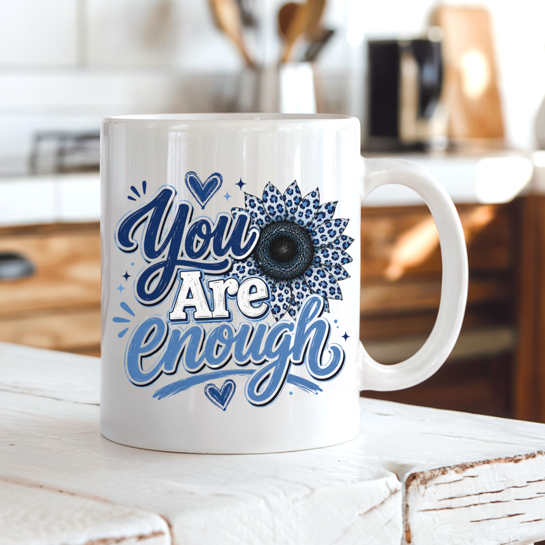 A white ceramic coffee mug with a blue and white graphic that reads 'You Are Enough' with a sunflower design, sitting on a rustic white wooden table in a blurred kitchen setting. Purchase yours from Coffee and Moo.