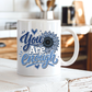 A white ceramic coffee mug with a blue and white graphic that reads 'You Are Enough' with a sunflower design, sitting on a rustic white wooden table in a blurred kitchen setting. Purchase yours from Coffee and Moo.