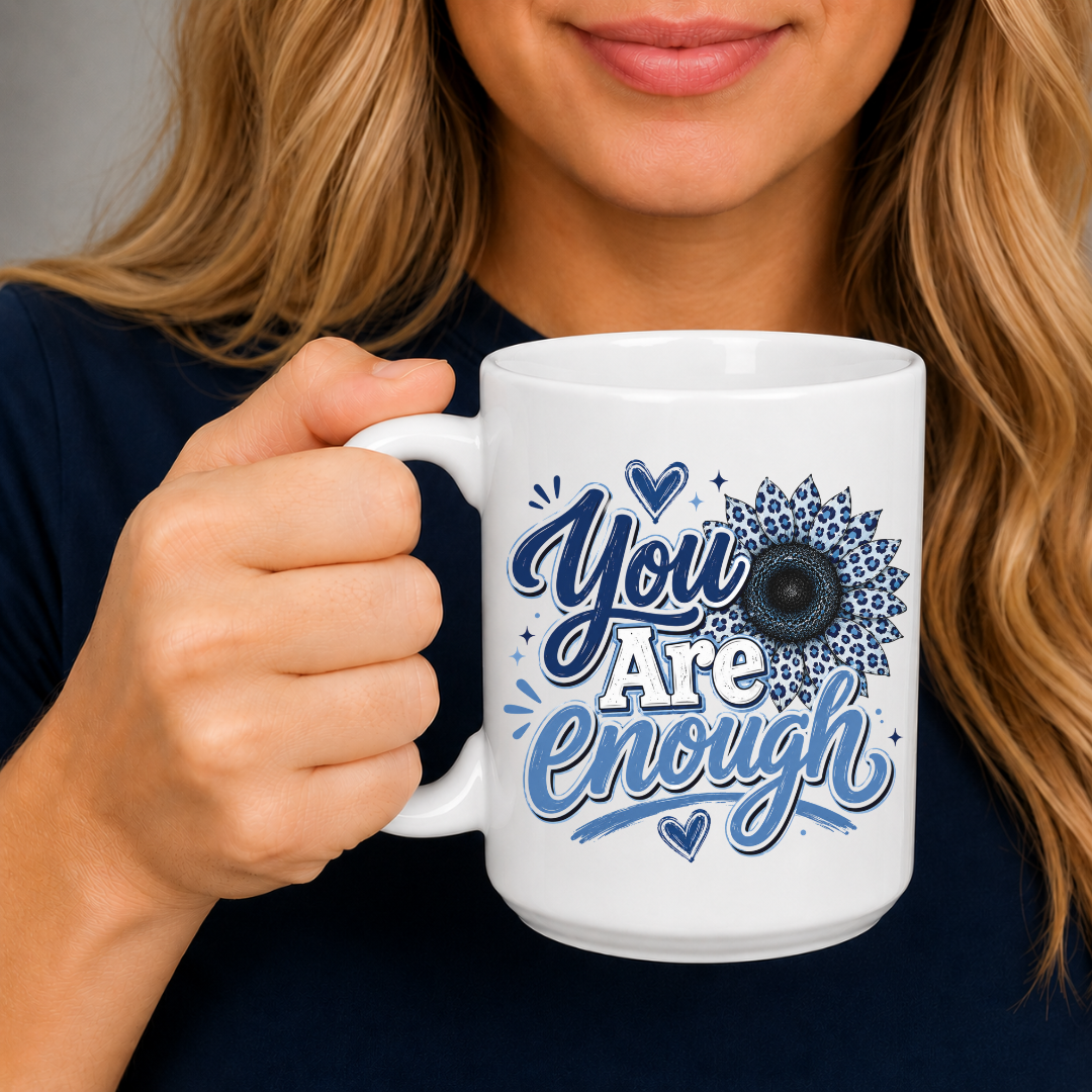 A woman holding a white ceramic mug with a blue graphic that reads 'You Are Enough' in stylized lettering with a sunflower design. Purchase yours from Coffee and Moo.
