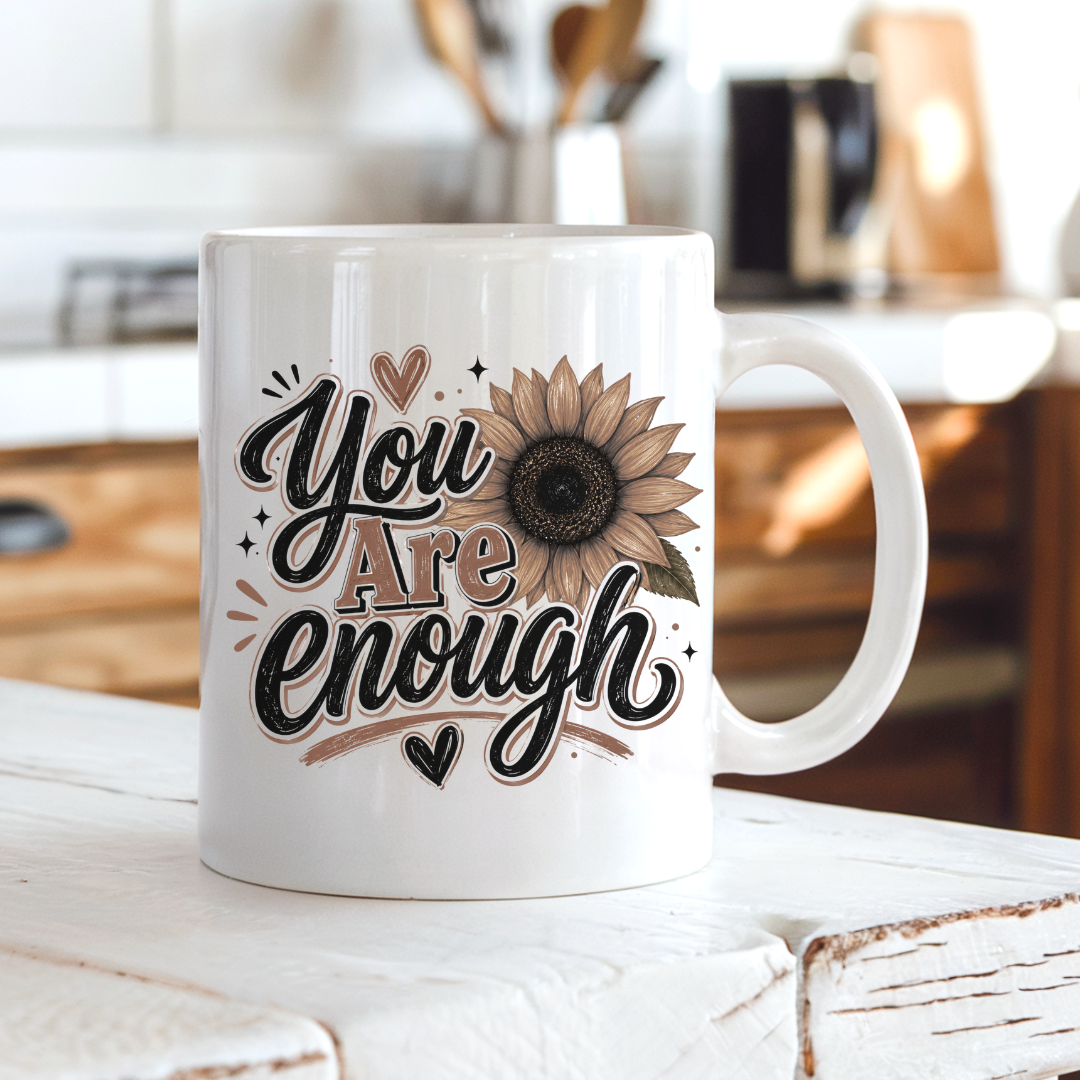 A white ceramic coffee mug with a brown and black 'You Are Enough' typographic design featuring a sunflower, resting on a rustic white wooden table in a warm, blurred kitchen setting. Purchase yours from Coffee and Moo.