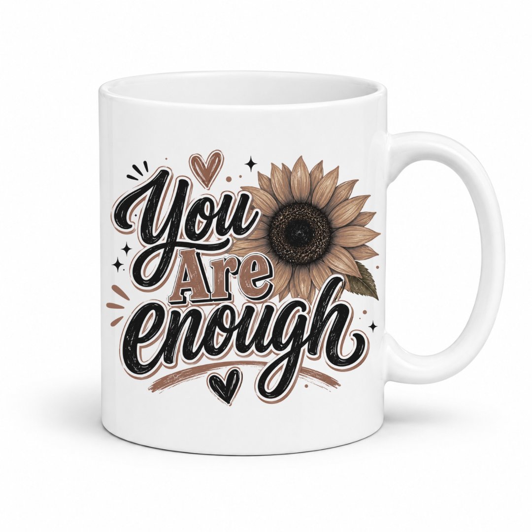A white ceramic mug featuring a printed design with the words 'You Are Enough' in black script and block lettering, accompanied by a brown sunflower and small hearts. Purchase yours from Coffee and Moo.