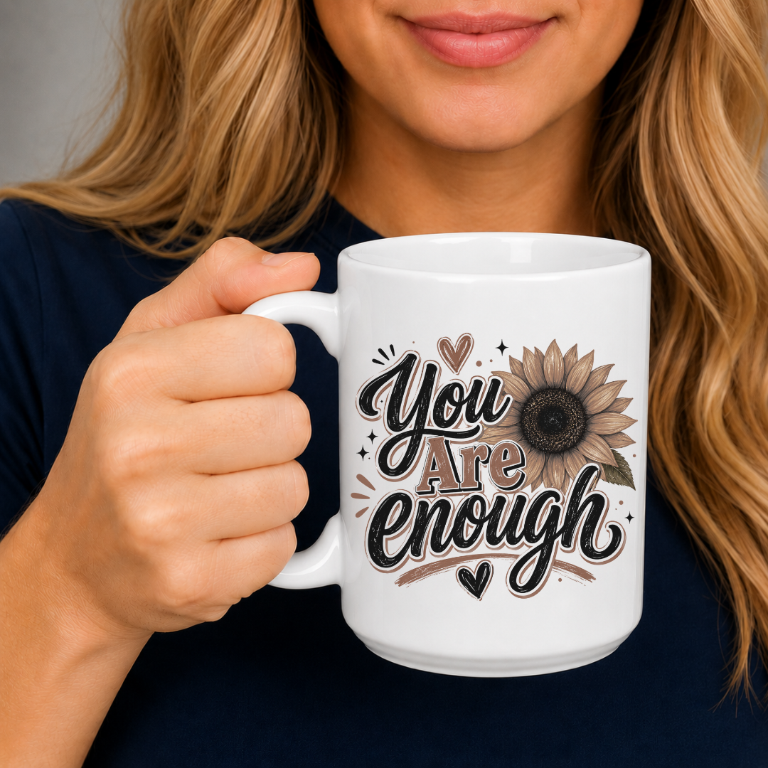 A person holding a white ceramic mug with a printed design that reads 'You Are Enough' in black script with brown and beige outlines, alongside a sunflower illustration. The person is wearing a dark navy top, and the background is a soft gray studio backdrop. Purchase yours from Coffee and Moo.