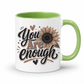 A white ceramic mug with a light green interior and handle, featuring a brown and black 'You Are Enough' typographic design featuring a brown sunflower, resting on a white background. Purchase yours from Coffee and Moo.