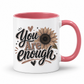 A white ceramic mug with a pink interior and handle, featuring a brown and black 'You Are Enough' typographic design featuring a brown sunflower, resting on a white background. Purchase yours from Coffee and Moo.