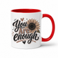 A white ceramic mug with a red handle and red interior, featuring a brown and black 'You Are Enough' typographic design featuring a brown sunflower, resting on a white background. Purchase yours from Coffee and Moo.